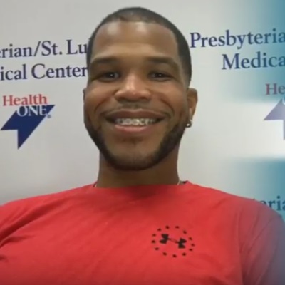 Marcus Bullock smiles while wearing an orange shirt and is standing in front of a banner with the Presbyterian/St. Luke's Medical Center logo.