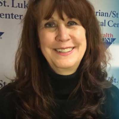 Jacquie Davey smiles while wearing a black turtleneck sweater and is standing in front of a banner with the Presbyterian/St. Luke's Medical Center logo.