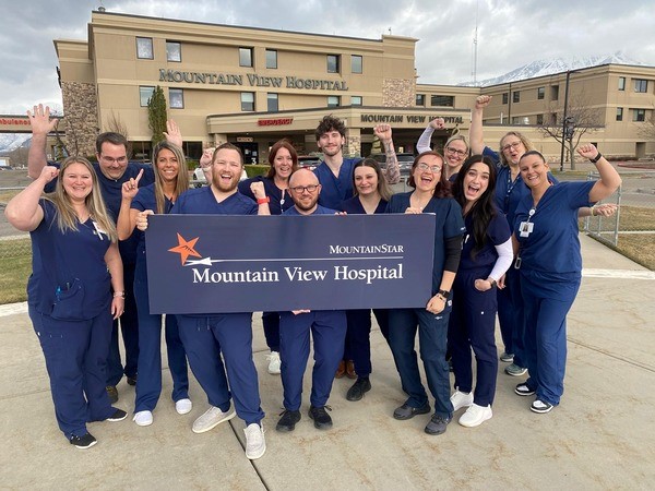 Colleagues who work in HCA Healthcare/MountainStar Healthcare's Mountain View Hospital ER smile and pose together outside the Payson, Utah facility after being awarded a Unit of Distinction. They're holding a sign that reads "Mountain View Hospital."