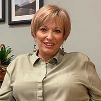 Sandee smiles while sitting in her office, wearing a tan button-up blouse.