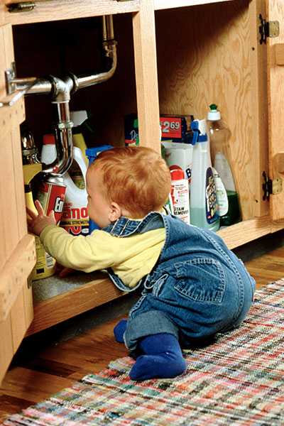 A baby sits on the kitchen floor, grabbing for household cleaning supplies under the kitchen sink.