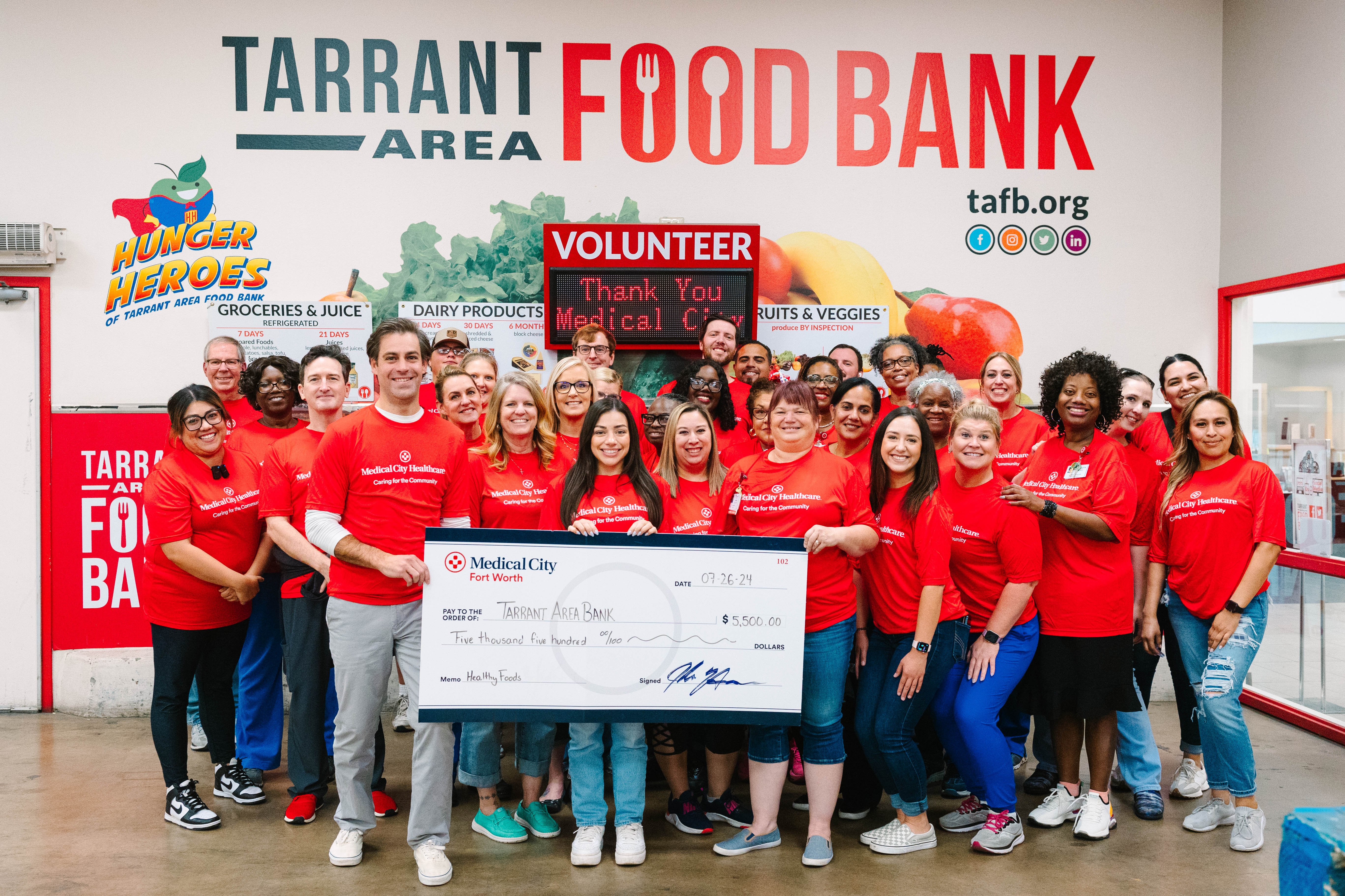 Food Bank volunteers in matching orange tee shirts pose with novelty sized $5,500 check.