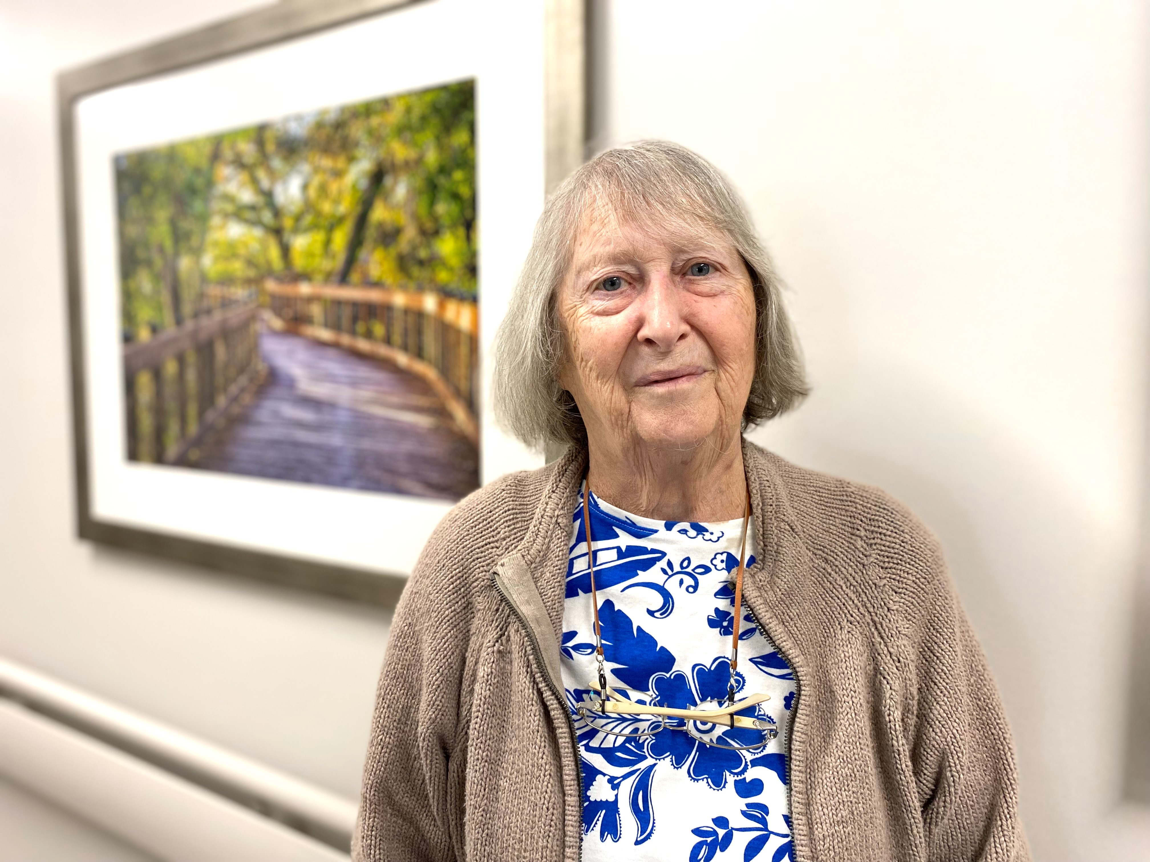 Patsy, recovered, standing in a hallway in front of a picture of a path through green woods.