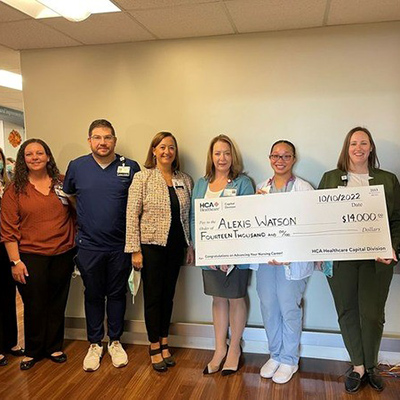 Alexis Watson stands with their coworkers while smiling and holding a large check for the scholarship they received from Johnson-Willis Hospital.