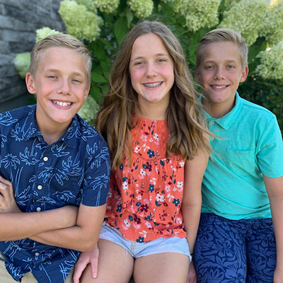 Carson, Alex, and Avery Kroeker smile while sitting next to flowers by their house.