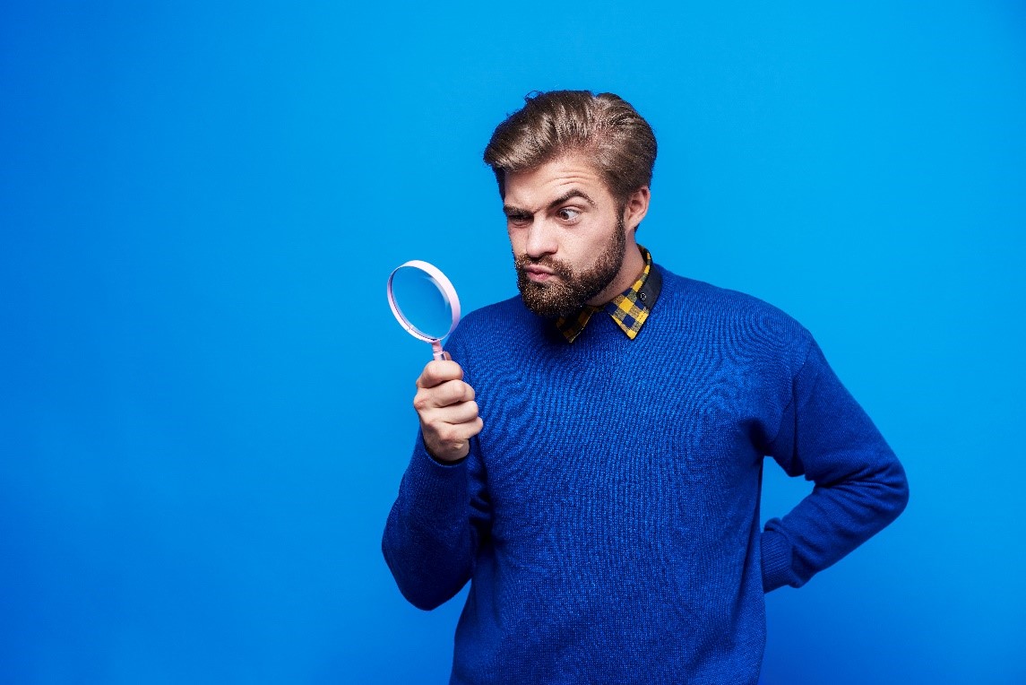 Man in a blue sweater, in front of a blue backgroung, looking through a magnifying glass.