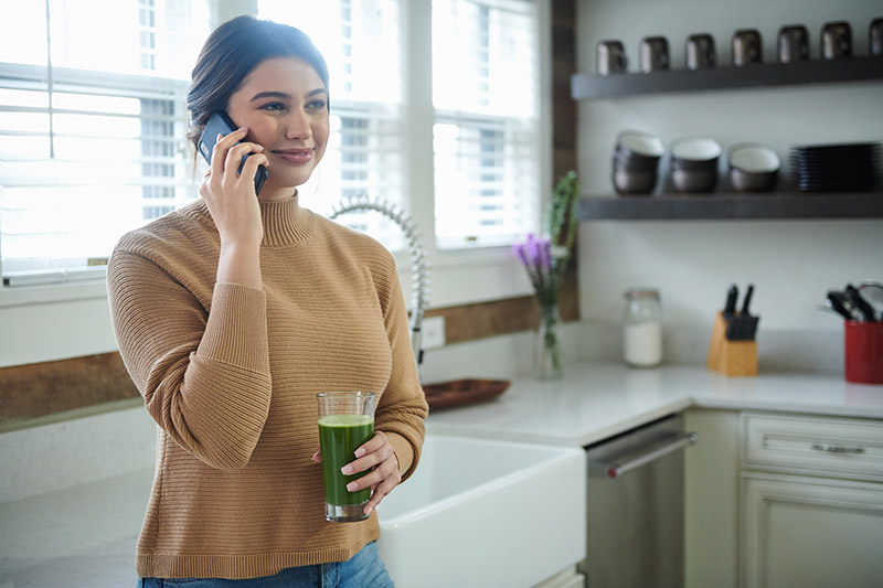 A light-skinned woman holds a cellphone in her right hand while standing in a daytime kitchen. In her left hand, she holds a clear glass with a green juice inside.