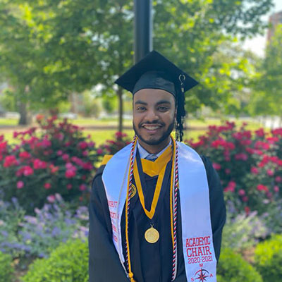 Mohamed Abdelrahman standing in front of a garden wearing his graduation cap and gown with his academic honor ropes around his neck.