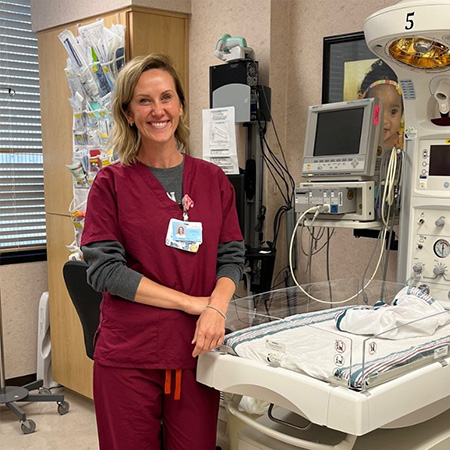 Cortney Parkinson smiles while wearing burgundy hospital scrubs, standing next to a newborn monitoring machine.
