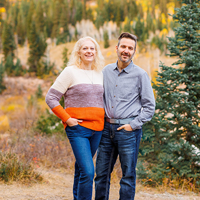 Michelle and Jason Kovalik smiling after their weight loss, standing together in nature, against a backdrop of pine trees.