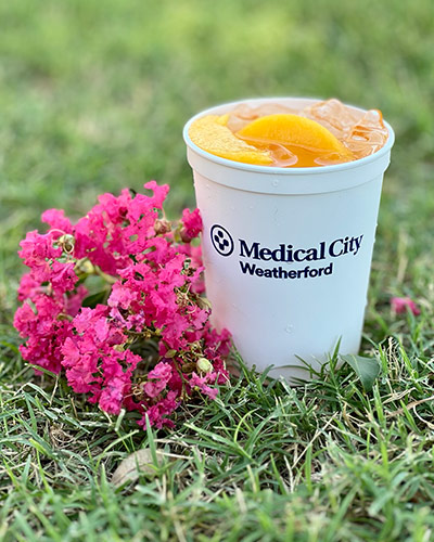 A cup of peach julep with the "Medical City Weatherford" logo on the cup, sitting in the grass with pink flowers sitting next to the cup.