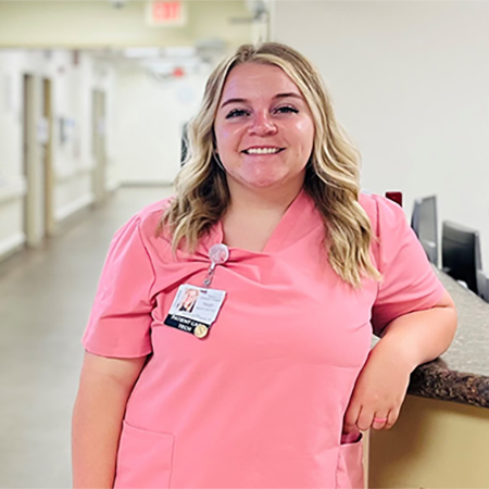 Teagan Beecher smiles while leaning on a nursing station counter, wearing pink hospital scrubs.