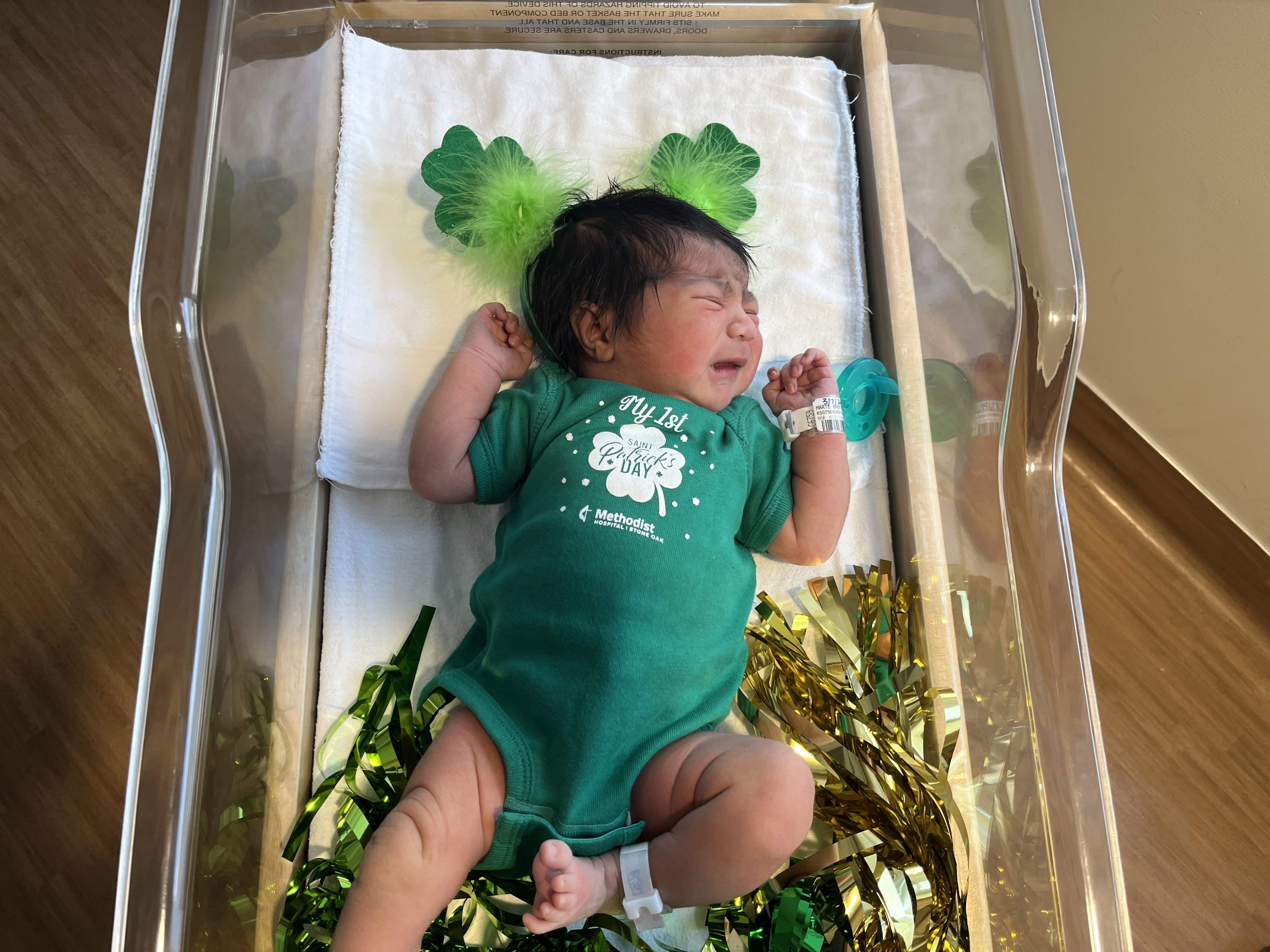 A baby in a green onesie lying in a hospital crib.