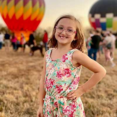 Audrey Sue smiles while wearing a floral print dress, hot air balloons in the background.