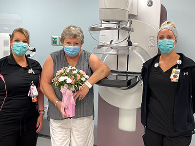 Mammographer Brandy Richards, Susan Schultz and Interim Director of Imaging Christine Trosper stand in front of the 3D mammography machine as Susan holds flowers. 