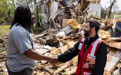 A volunteer shakes a Red Cross worker in front of the debris of a collapsed building in a wooded setting.