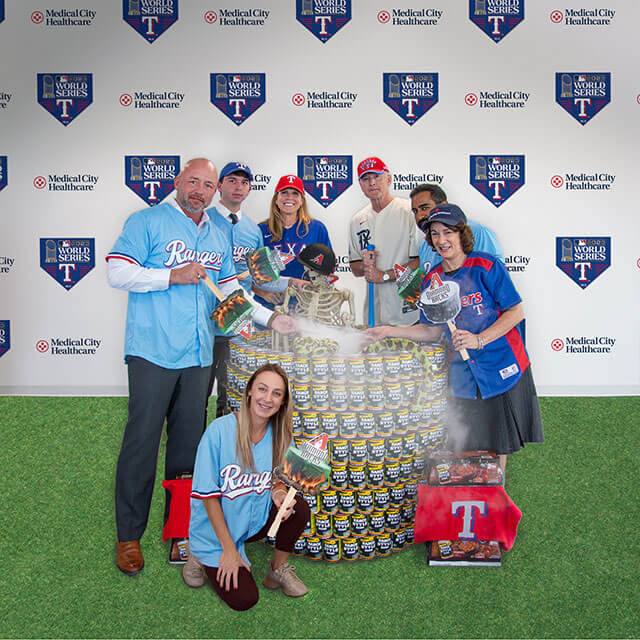 A group of hospital staff smile while standing with the "Witching For a Rangers World Series Win" can sculpture during a Halloween-themed food drive.
