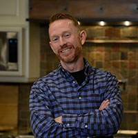 Ben Gowans smiles and is crossing his arms, while standing in his kitchen wearing a blue and black plaid button-up shirt.