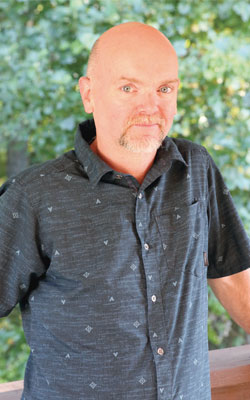 Bobby wears a patterned grey button-up shirt and is standing next to a tree.