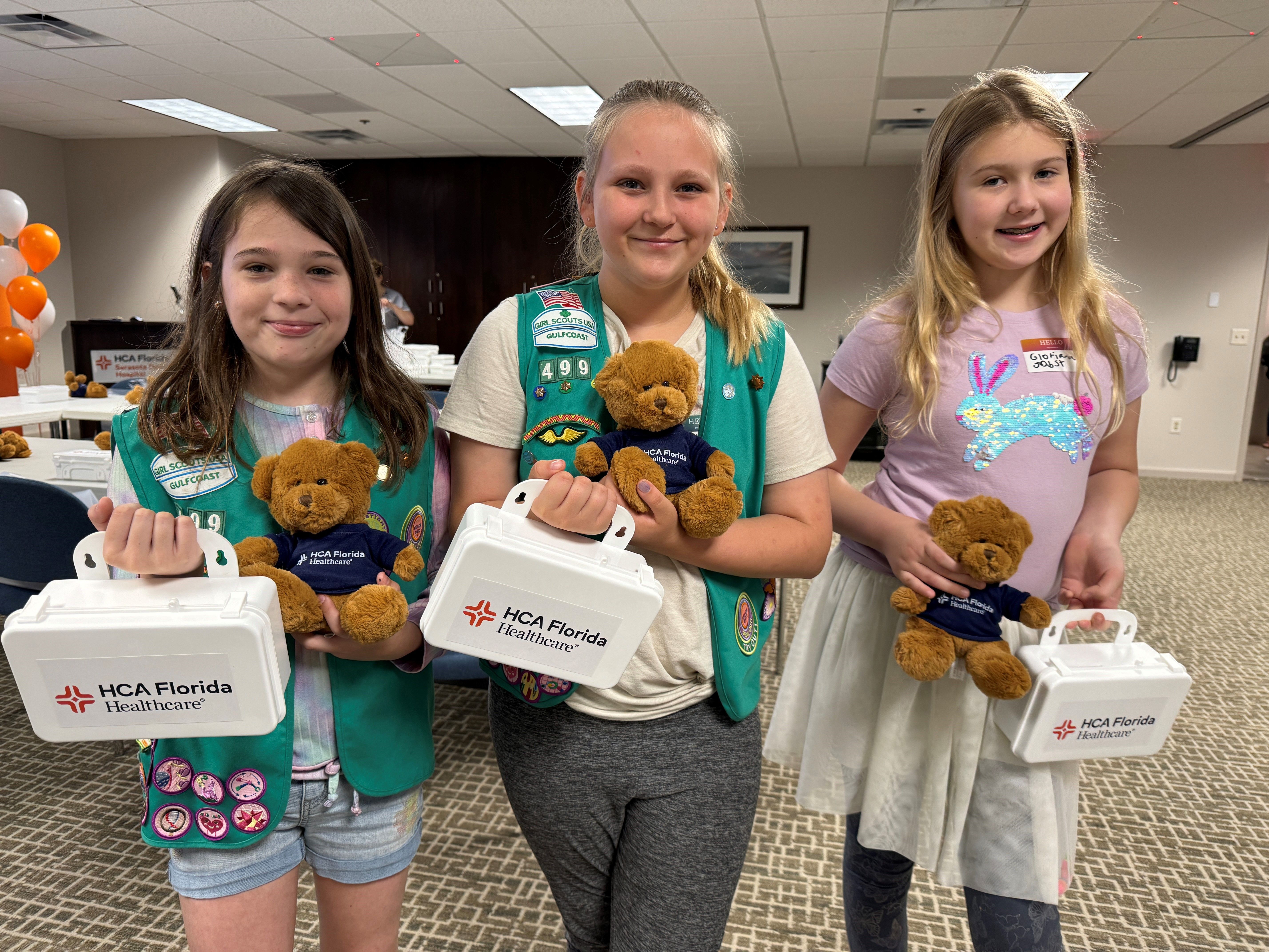 Three girl scouts stand smiling with first aid kits and teddy bears