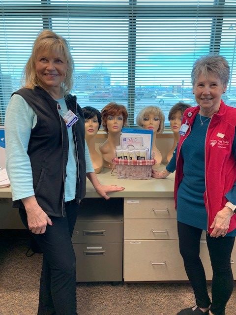 Two Sky Ridge Medical Center volunteers stand next to a desk displaying mannequin heads with various wig colors and a care basket.