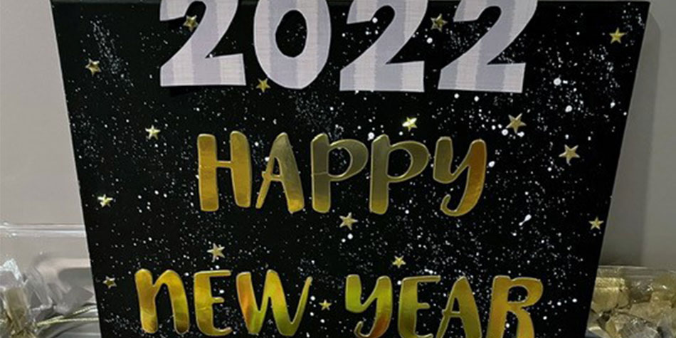 A handmade sign with a decorative night sky background reads in overlaid bold white and gold lettering, " 2022 Happy New Year."