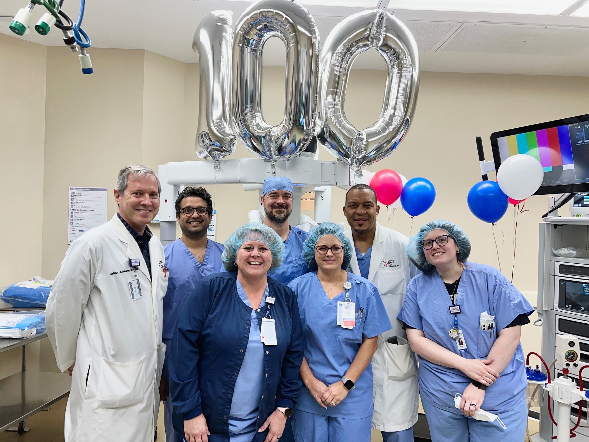 Robotic Surgeons and staff pose in operating room in front of "100" balloons.