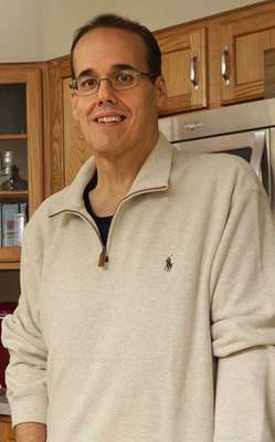 Todd smiles while wearing a beige pullover and standing in his kitchen.