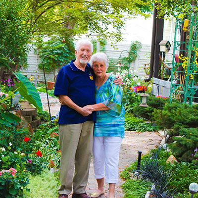 John and Wanda Blankenstein are standing together hand in hand on a path surrounded by a vibrant tropical plants, trees, and lawn ornaments