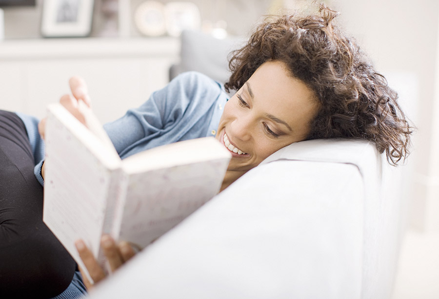A medium-light skinned toned person with shoulder length curly brown hair and denim blue shirt is curled up on a couch reading a white-colored book.