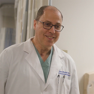 Dr. Mizrahi smiles while walking in the hospital, wearing a white scrub jacket.