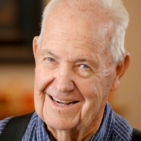 Jerry Olsen smiles while wearing a blue and white plaid button-up shirt and suspenders.