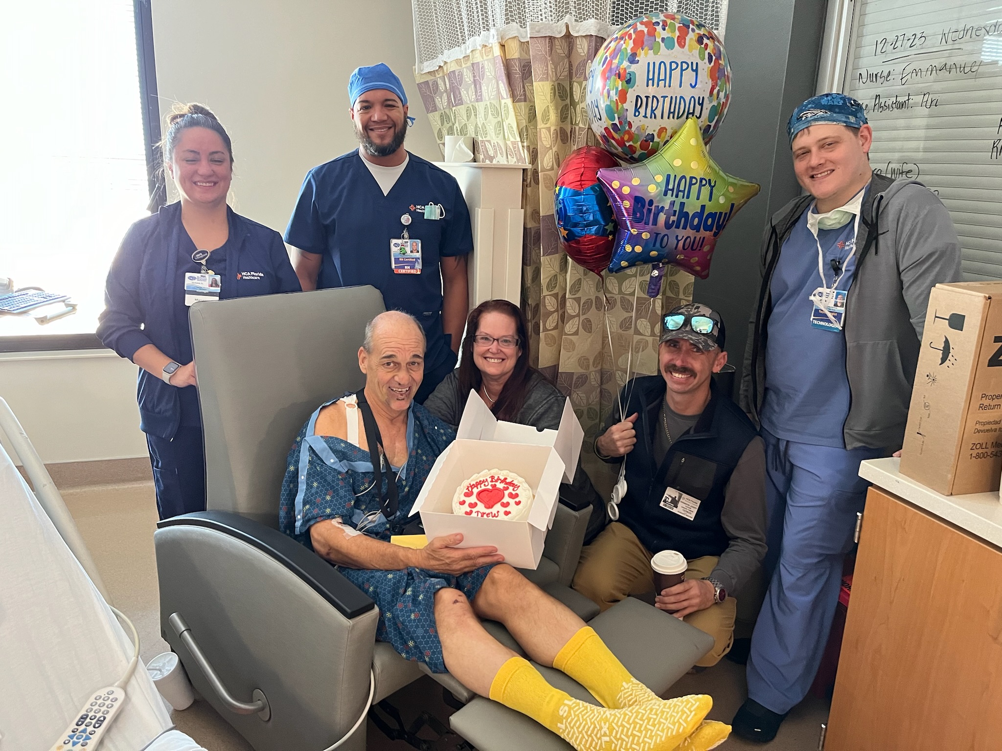 Drew sitting with a cake and a balloon, surrounded by his heroes, the colleagues and EMS team celebrating his birthday after saving his life.
