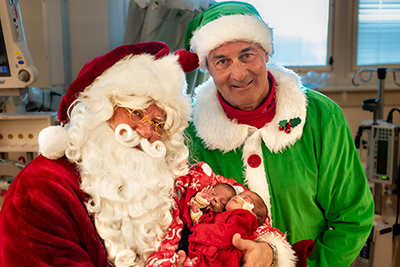 Rob Holcomb, a doctor at Overland Park Regional Medical Center dressed as Santa, smiles while holding two babies in the NICU, while an elf stands behind him.