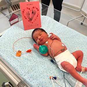 A newborn baby lies in a bed with a Valentine's card