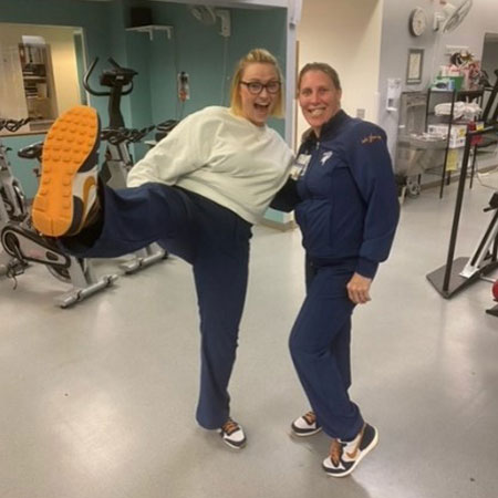 Clinical exercise physiologists' Alicia Satterwhite and Lacey Acree pose in a room with exercise equipment, as Satterwhite extends her right leg in the air