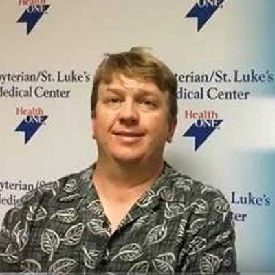 Paul Pursell smiles while wearing an olive green botanical print button-up shirt, and is sitting in front of a banner with the Presbyterian/St. Luke's Medical Center logo.