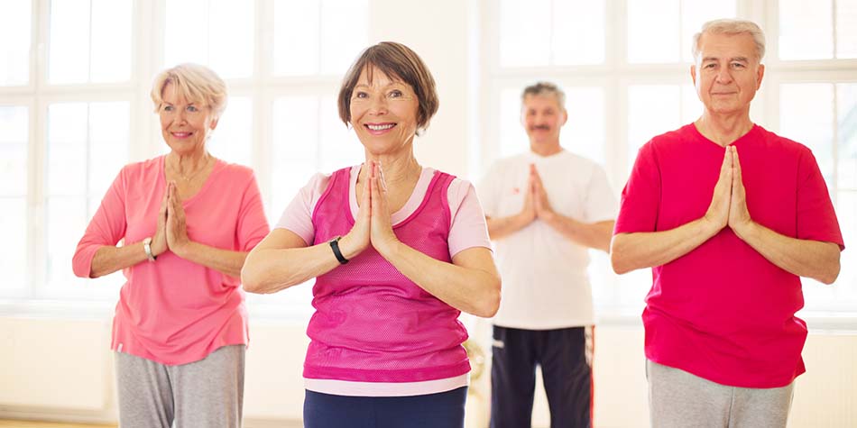 A group of retiree's hold a standing pose with their hands together during a yoga session.