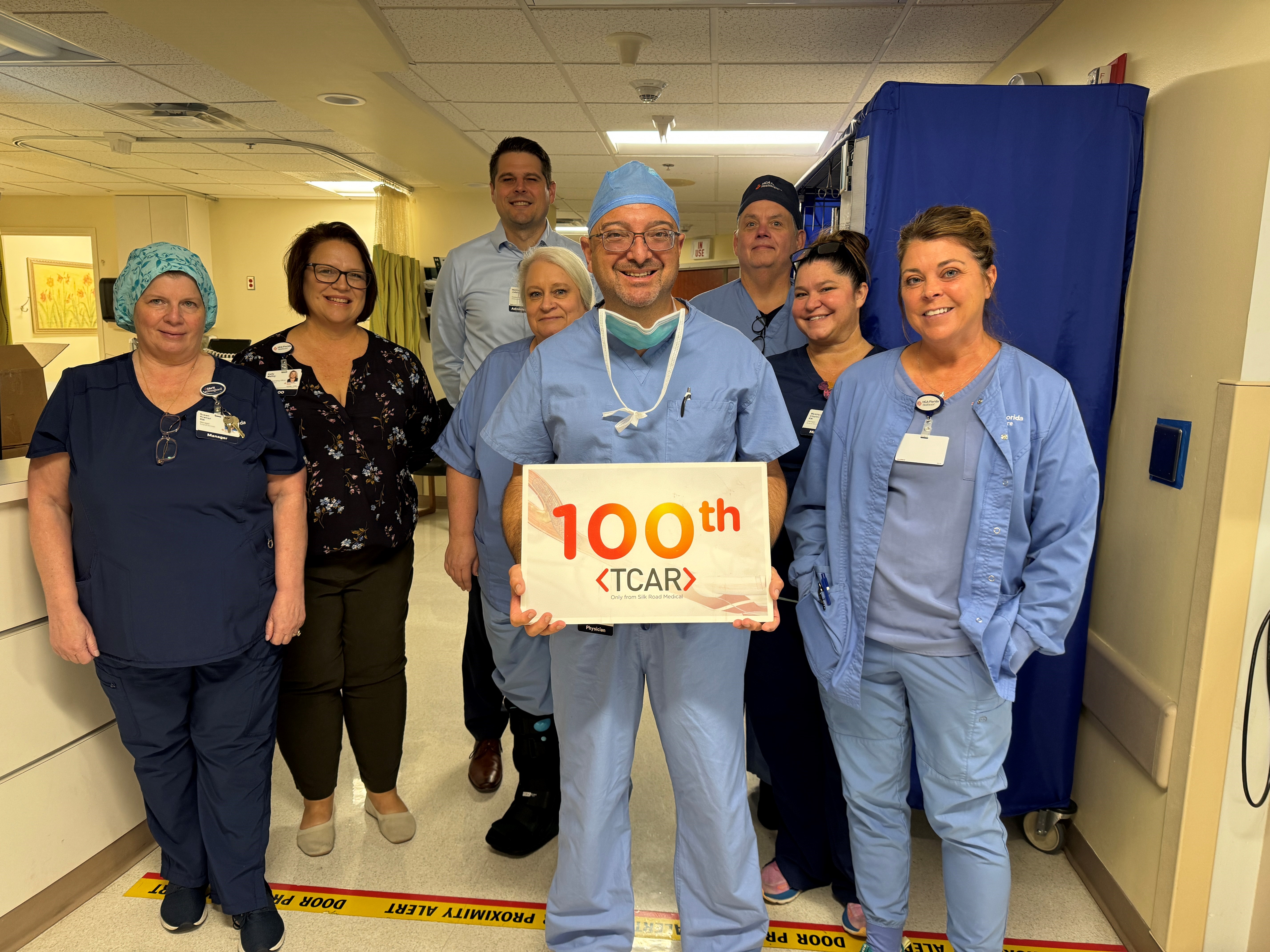 Doctor stands with surgical team smiling and holding sign that reads 100th.