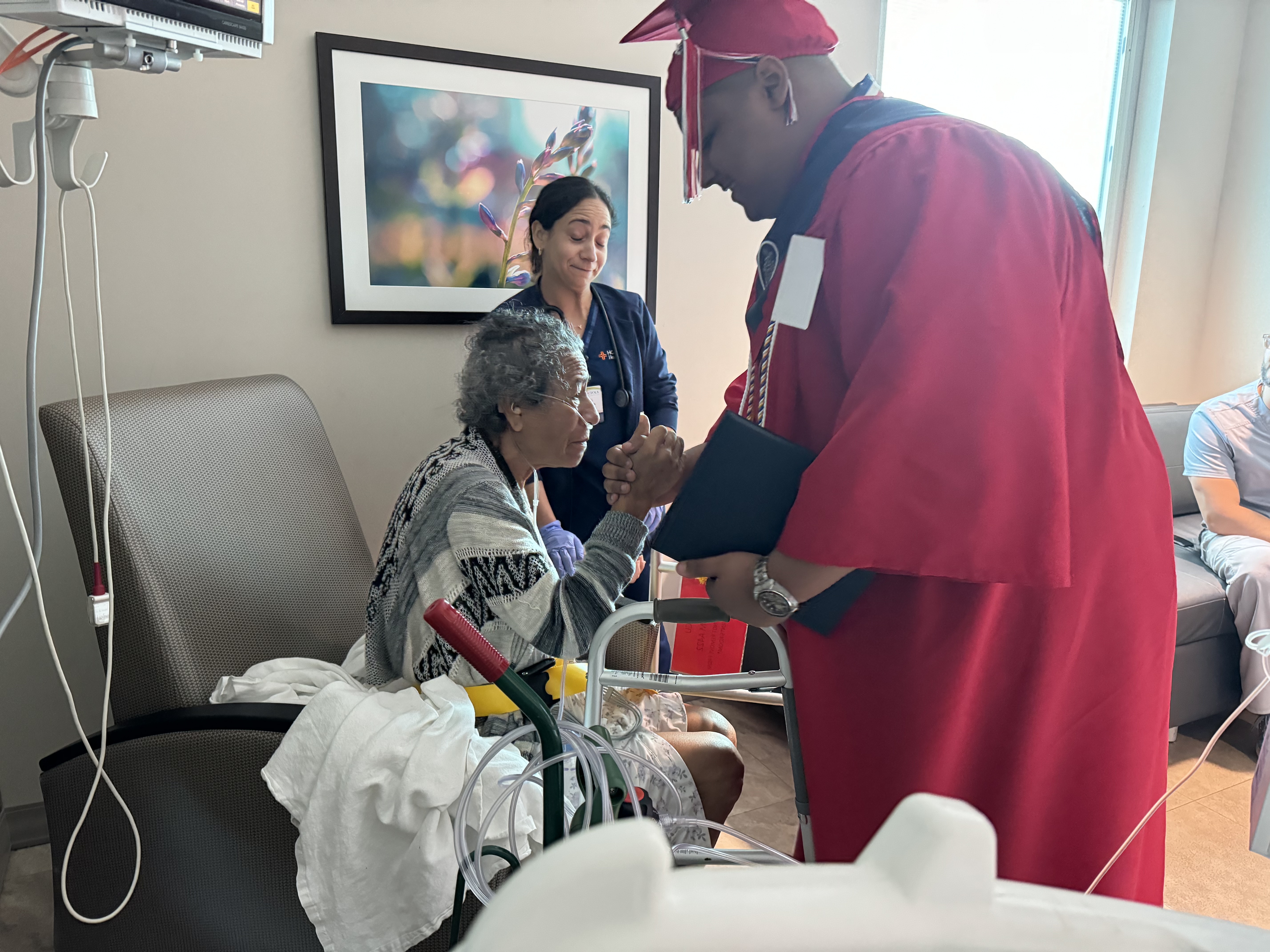 High school graduate in red robe and cap with grandmother and nurse in hospital room
