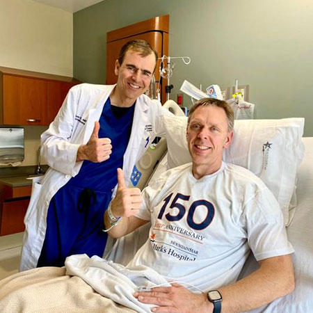 Tom Dokken on his hospital bed, wearing a St. Mark’s Hospitals 150th ablation t-shirt, giving a thumbs up with Dr. Day standing beside him.
