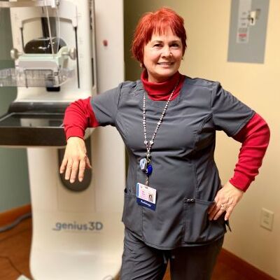 Liz Zenter smiling, wearing a  red turtleneck under her dark blue radiology uniform, in front of a Genius 3D mammography exam machine. 