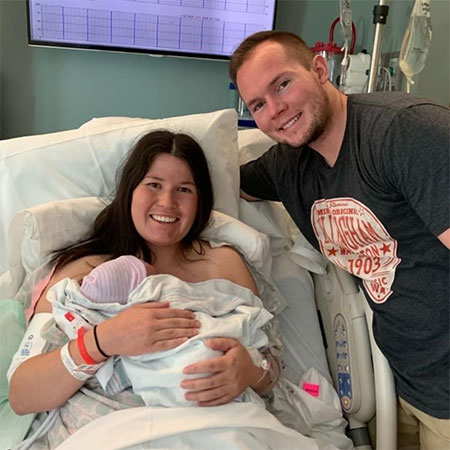 Haeli sitting upright on her hospital bed, holding her newborn baby Spencer, with her husband Carlyn by her side.