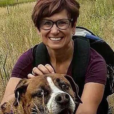 Joyce Hanson smiles while hugging her dog on a hiking trail.