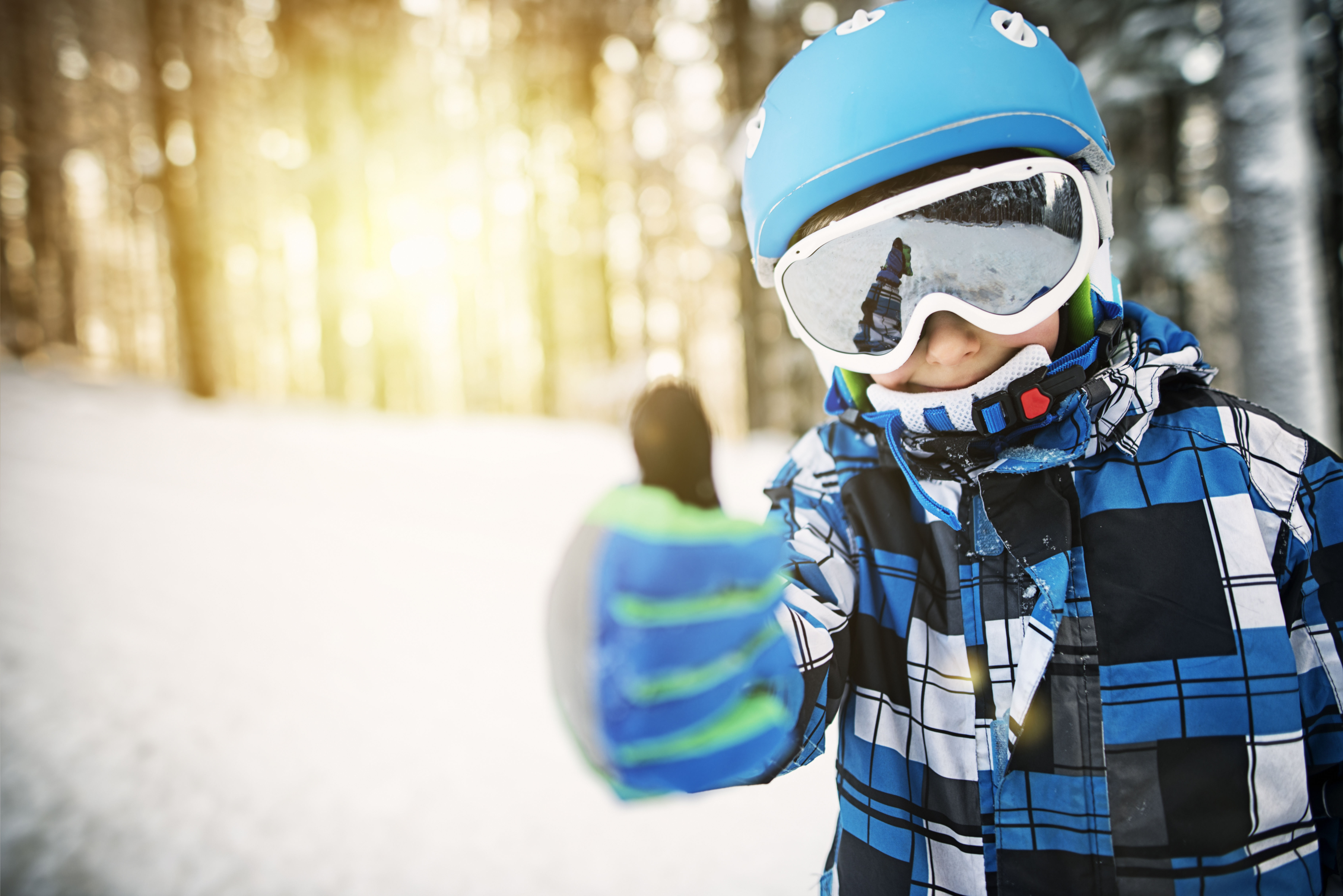 Child in blue helmet and white ski gogles gives a thumbs up in a gloved hand.