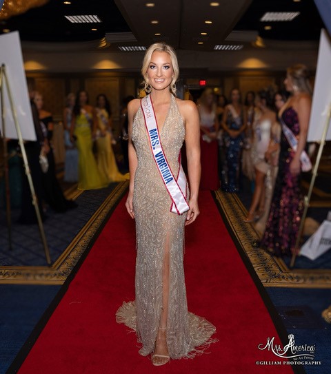 Amanda Miller walks a read carpet wearing her red white and blue colored "Mrs. Virginia" pageant sash across a sequin dress.