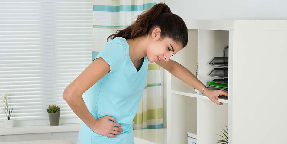 A woman bends over in pain while holding her stomach and bracing herself with her left arm against a bookshelf.