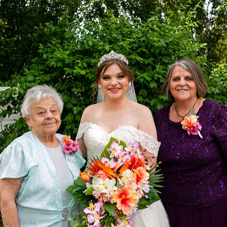 Pam Paulsen stands in between family members while wearing a white wedding dress and holding a floral bouquet in an outdoor setting