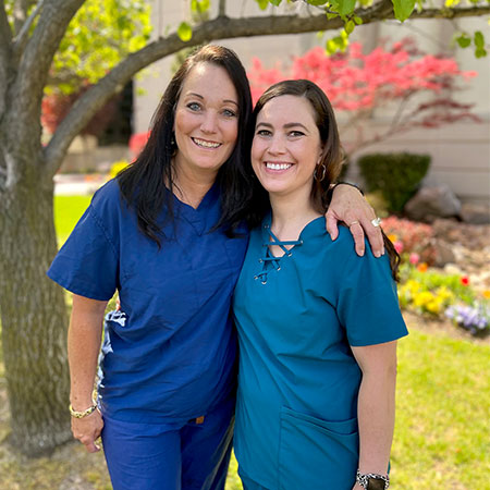 Marilyn Allred has her arm rested on her daughter Mikayla Allred as they pose in nursing scrubs near some trees and grass.