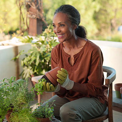 A medium-skinned woman with tied-back black and grey holds a potted plant in her gloved hands. She is seated in an outdoor patio with other nearby plants, where there is warm sunny tree-line behind her. 